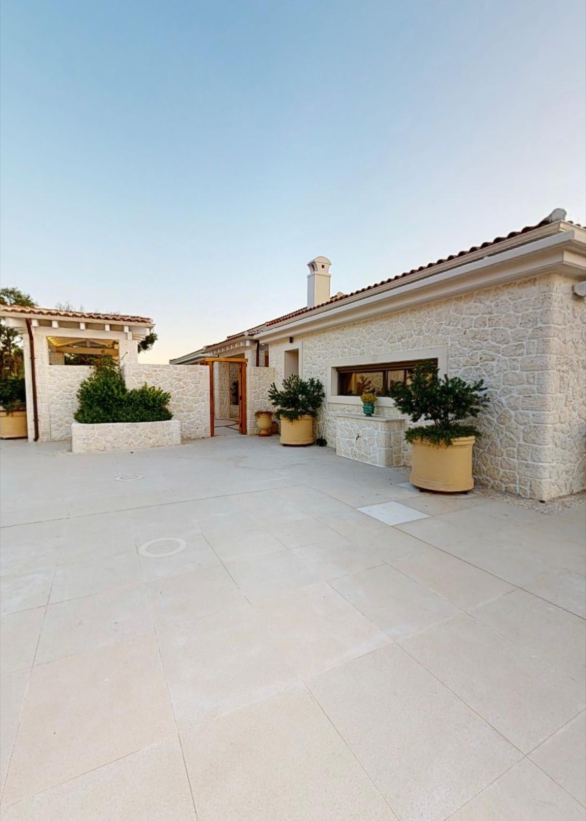 White rough-cut polygonal stone cladding, terracotta roof with stone chimney, sand-toned stone paving courtyard, large terracotta planters, pergola at left