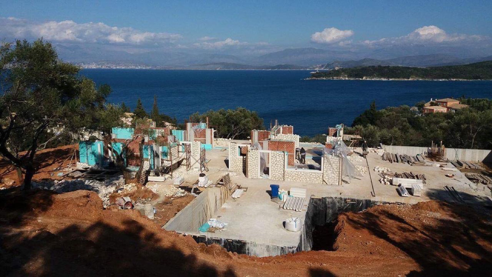Rising walls of brick and turquoise block being clad in white stone, raw excavated earth and pool shell in foreground, full bay, islands and mainland mountains 