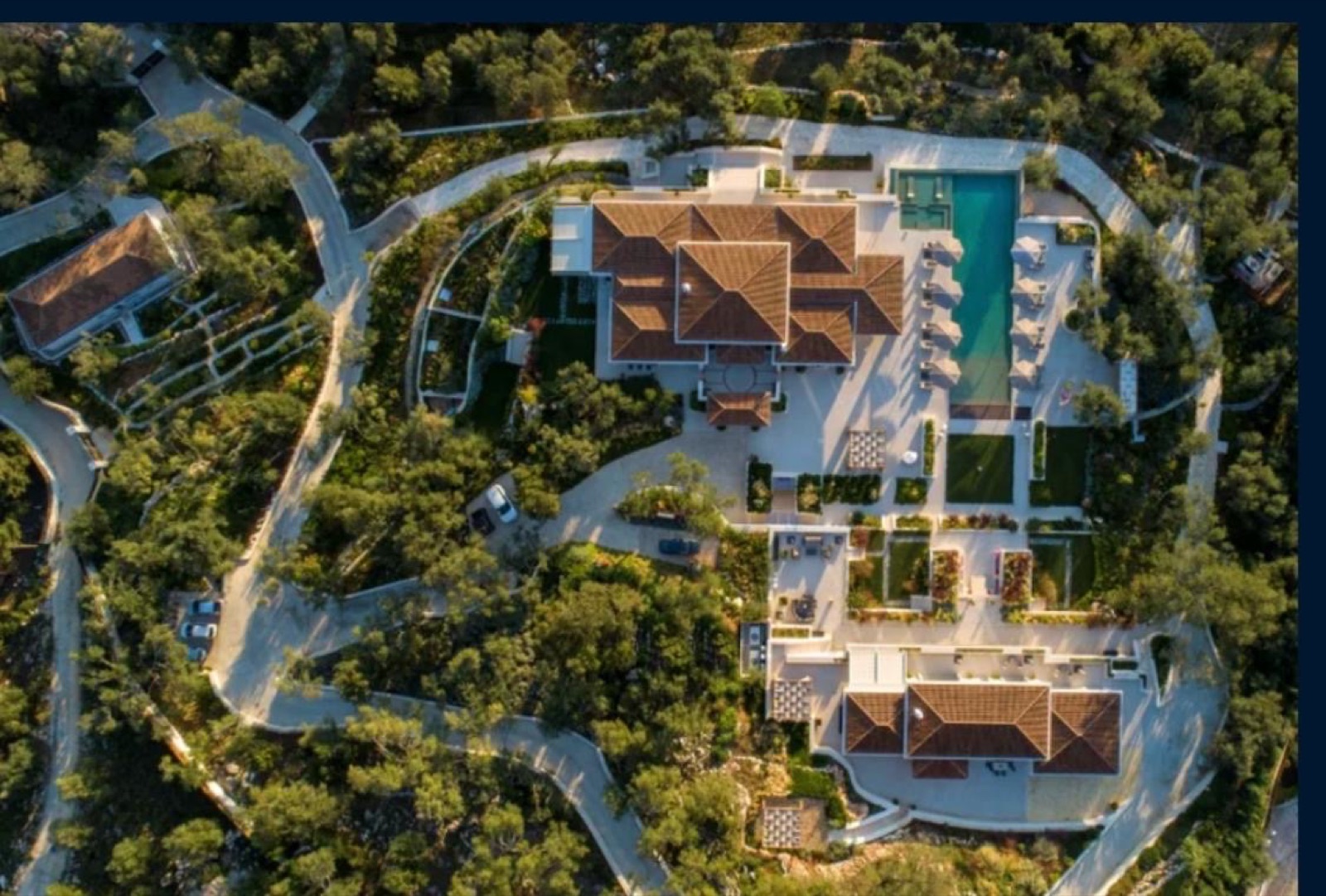 Tiled roofs of main villa and guest building, pool with loungers, lawns, and winding white drives threading through olive groves