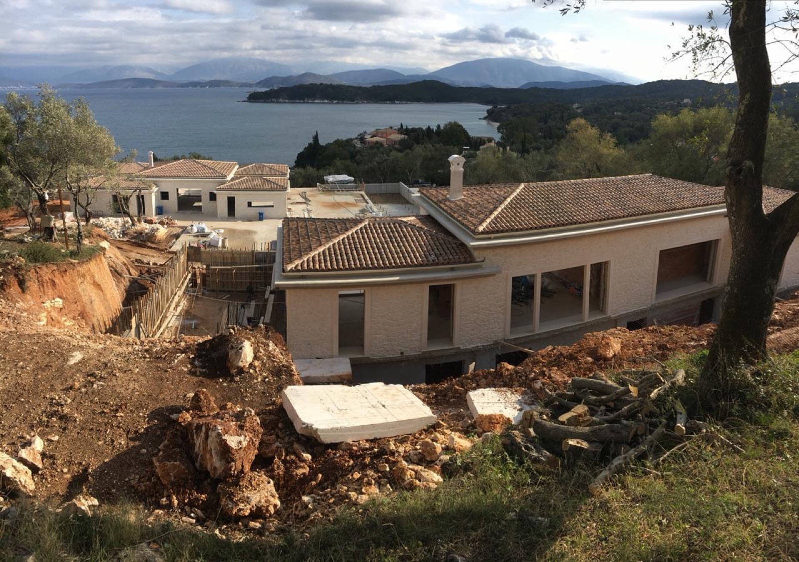 Villa shell with terracotta roof and render complete, raw earthworks and timber formwork in foreground, sweeping view over the bay, headlands and mountains unde