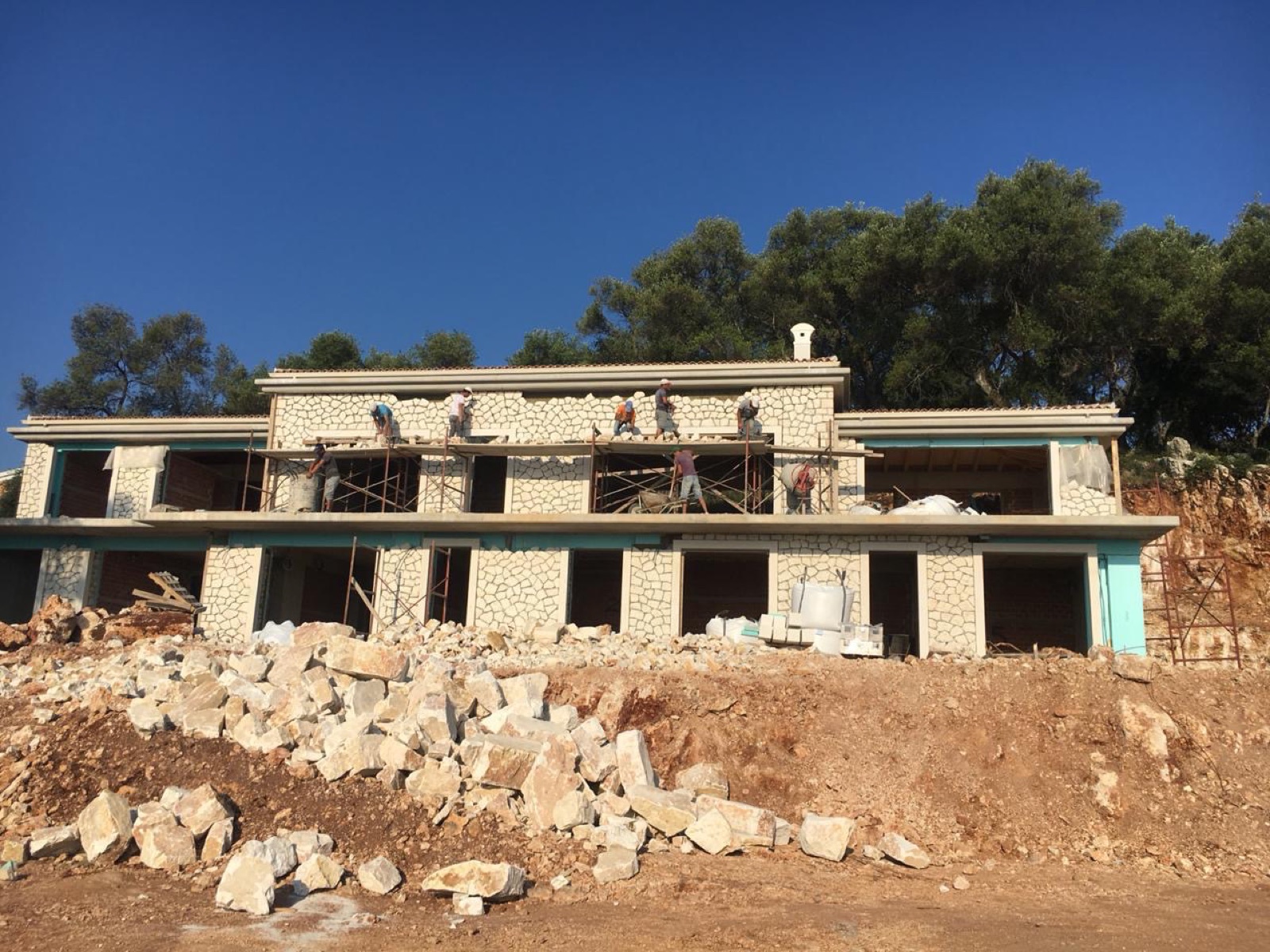 A crew on scaffolding points the white polygonal stone cladding across both storeys, quarried stone blocks in the foreground.