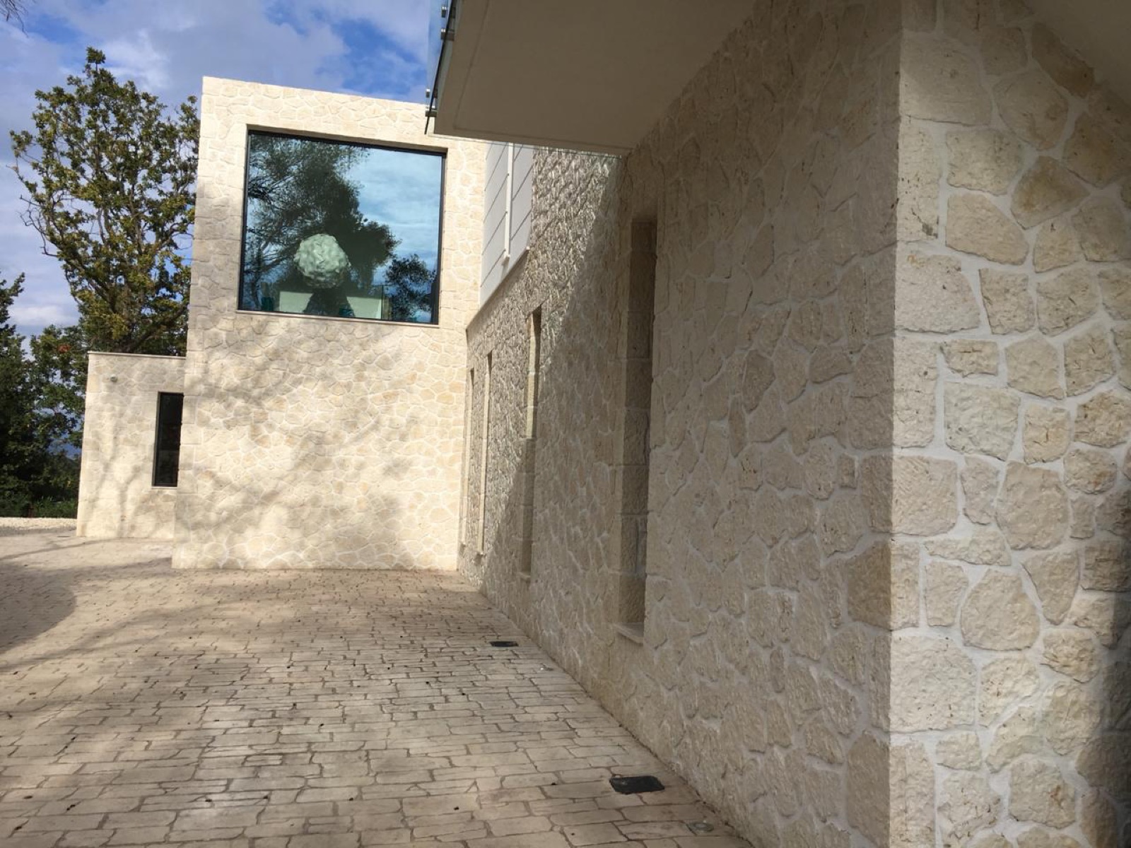 Stone-clad volumes with a large square picture window reflecting trees and sky, stone-paved forecourt, blue sky with scattered clouds