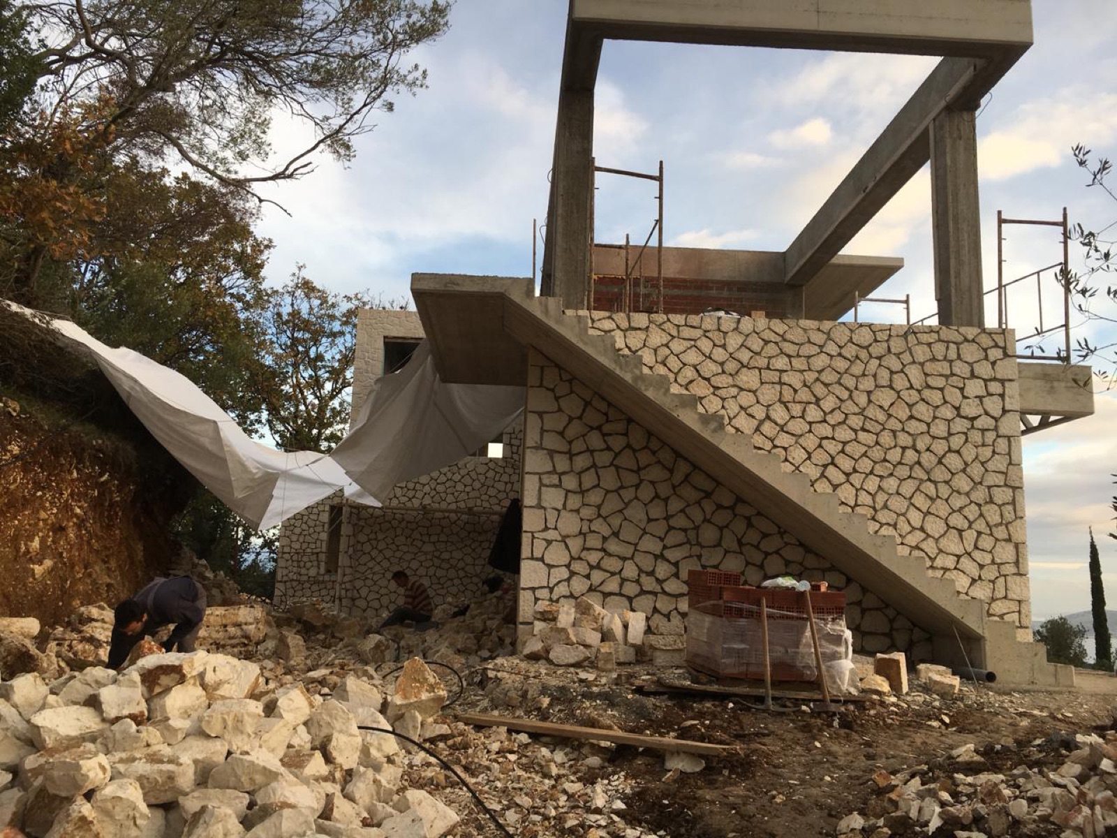 Stone cladding being applied to the external staircase wall, scaffolding around the concrete frame and masons among the stones.
