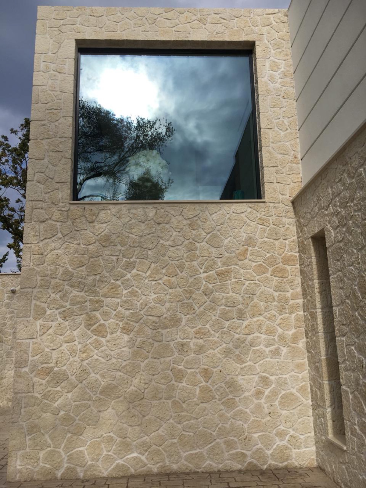 Large dark glass square reflecting dramatic clouds and an olive tree, surrounded by cream polygonal limestone with pale mortar joints