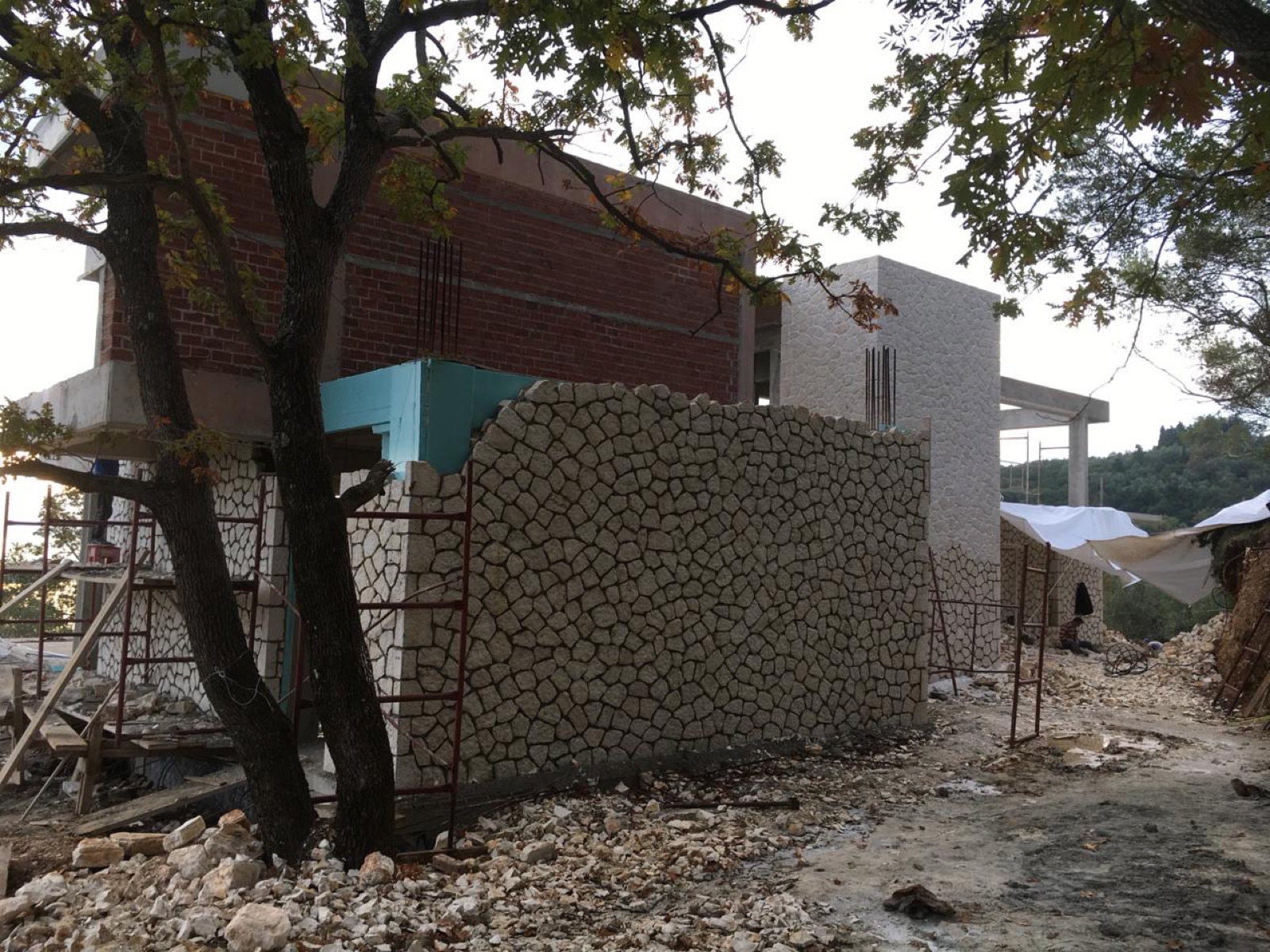 The stone boundary wall part-built at dusk, scaffolding on the upper storey and an oak tree in the foreground.