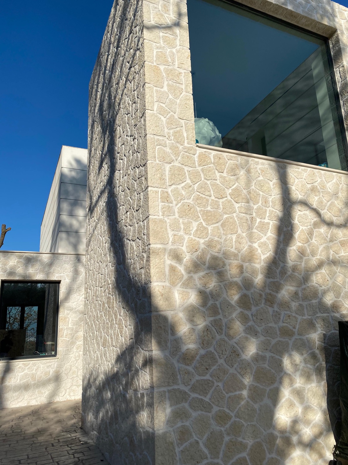 Stone-clad volume with glass corner window above, dappled tree-branch shadows raking across the limestone, cobbled paving below