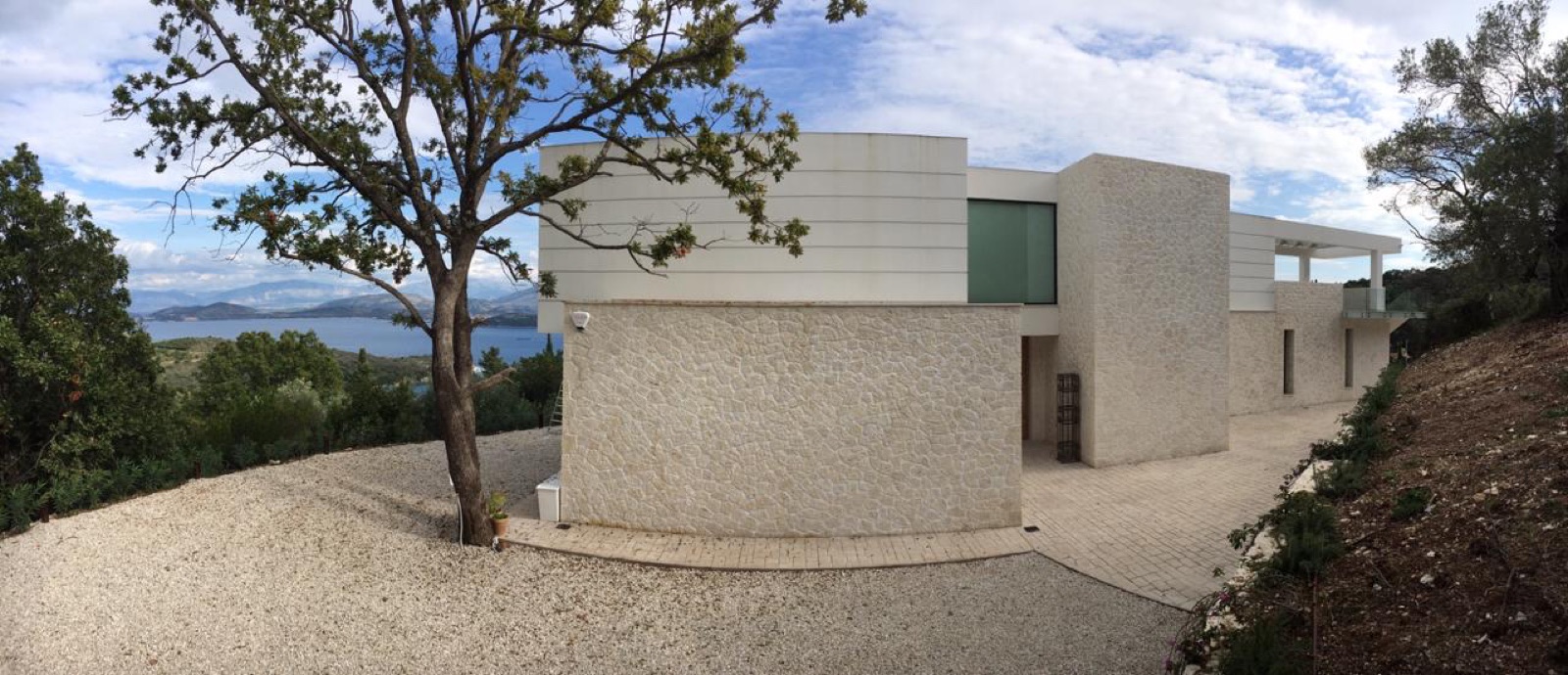 White modern volumes over curved stone-clad base walls, gravel forecourt with lone oak, sea and mainland mountains behind