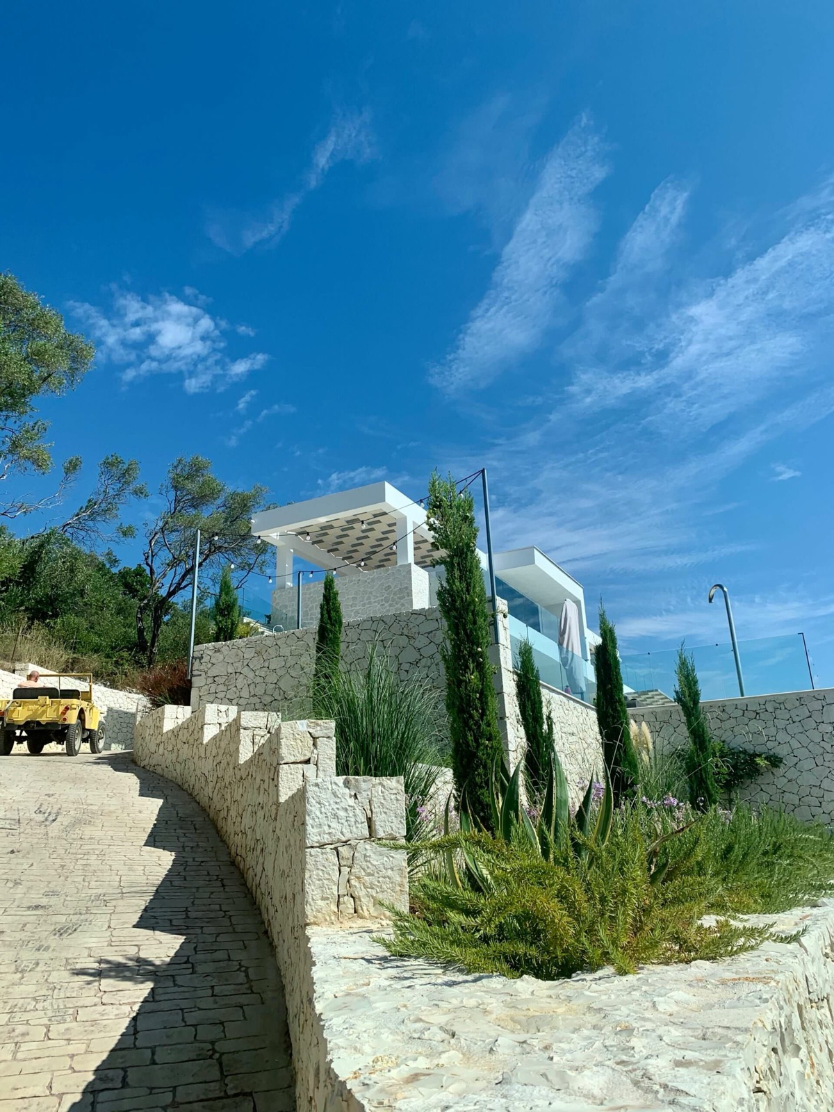 Looking up the stone-paved driveway between curved limestone retaining walls, with cypresses and white pergola terraces above.