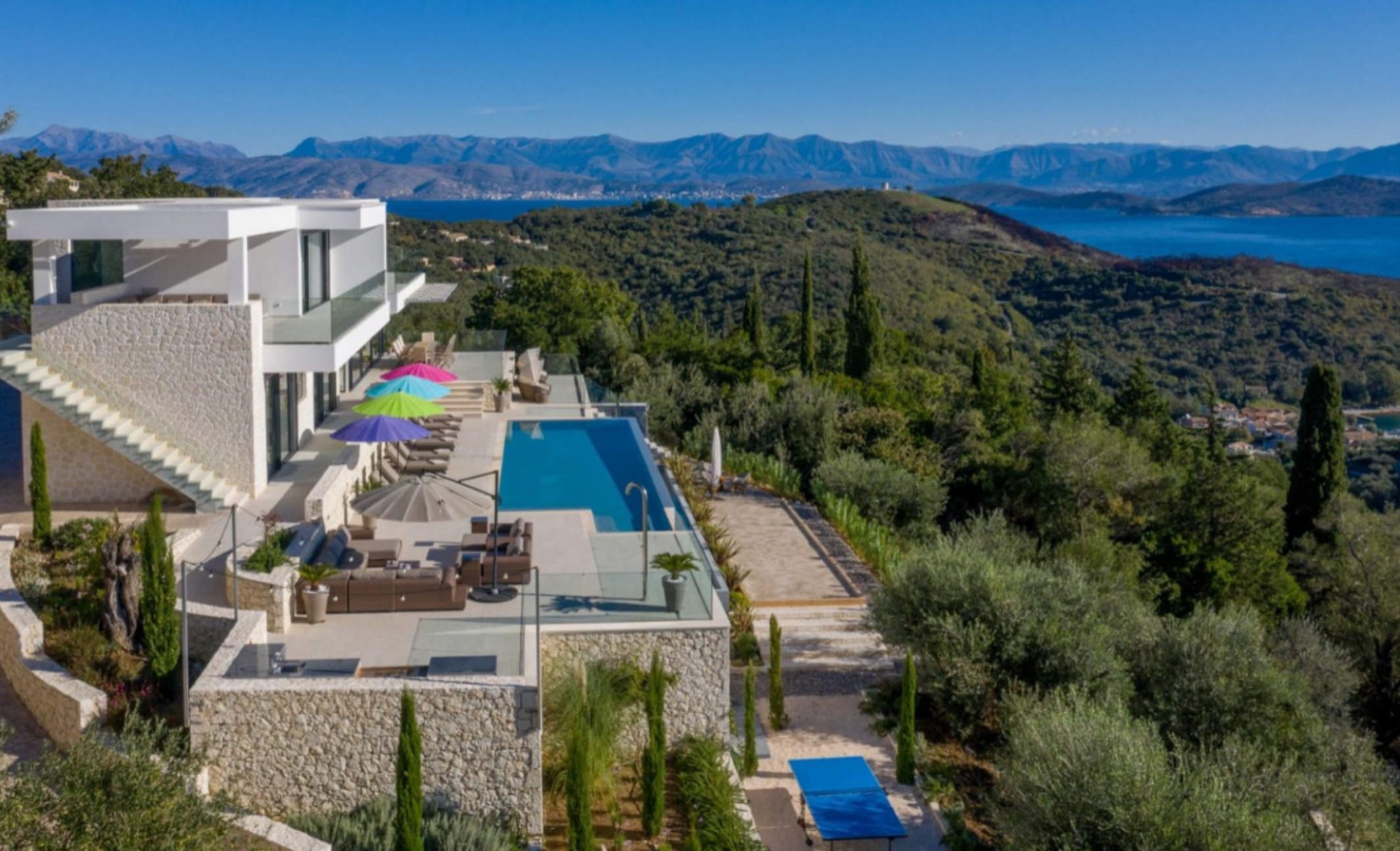 Infinity pool, stone retaining walls and terraces stepping down the hillside, loungers and umbrellas, sweeping Ionian sea, islands and mountain backdrop