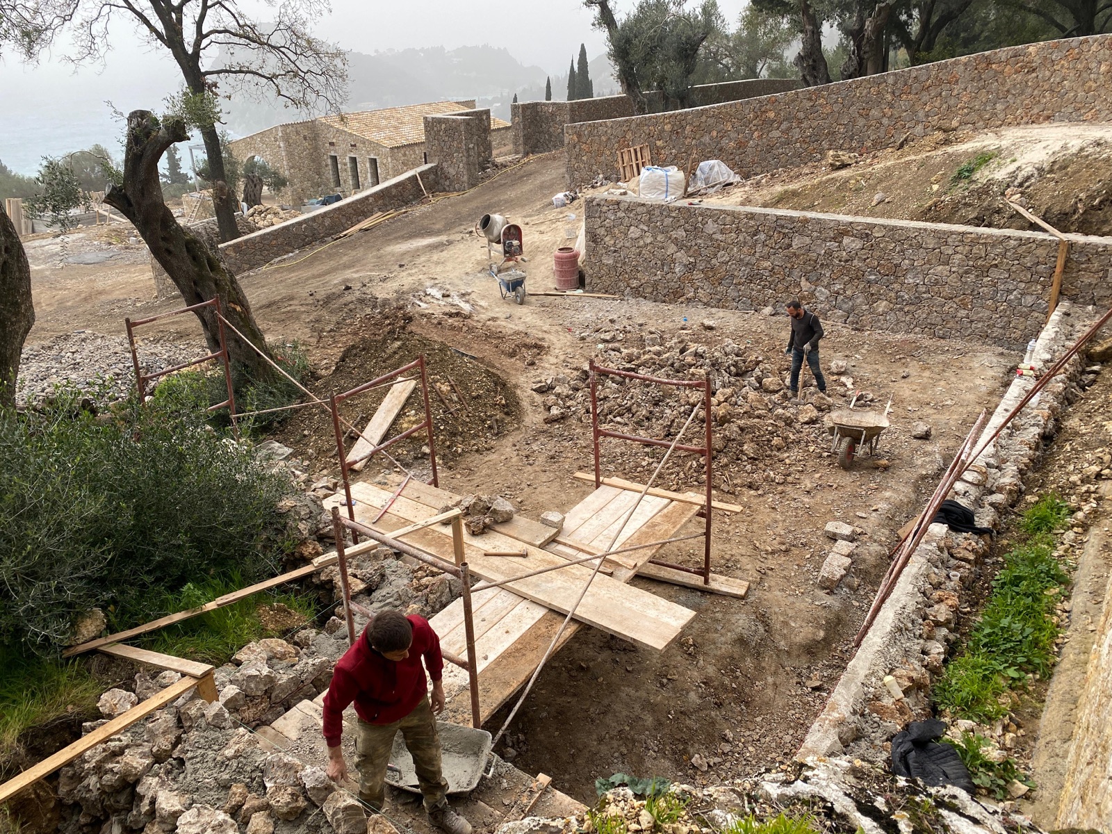 Scaffolding and timber walkways between rising stone retaining walls, the finished perimeter wall and misty hills behind.