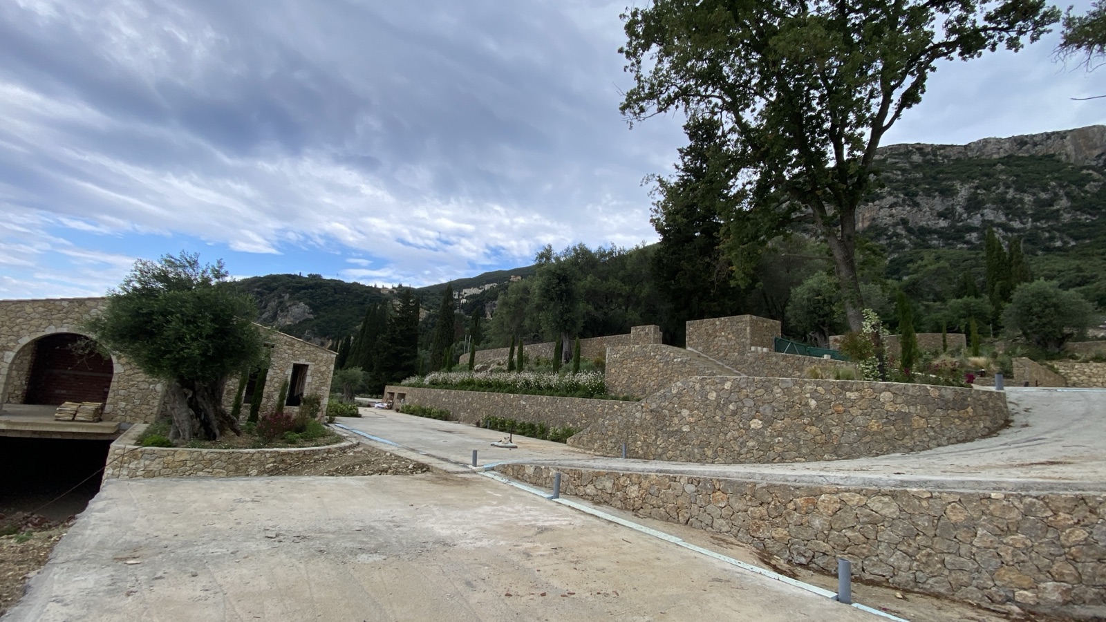 Curved battered stone retaining walls we built beneath a limestone cliff, with an arched stone outbuilding and a mature olive tree.