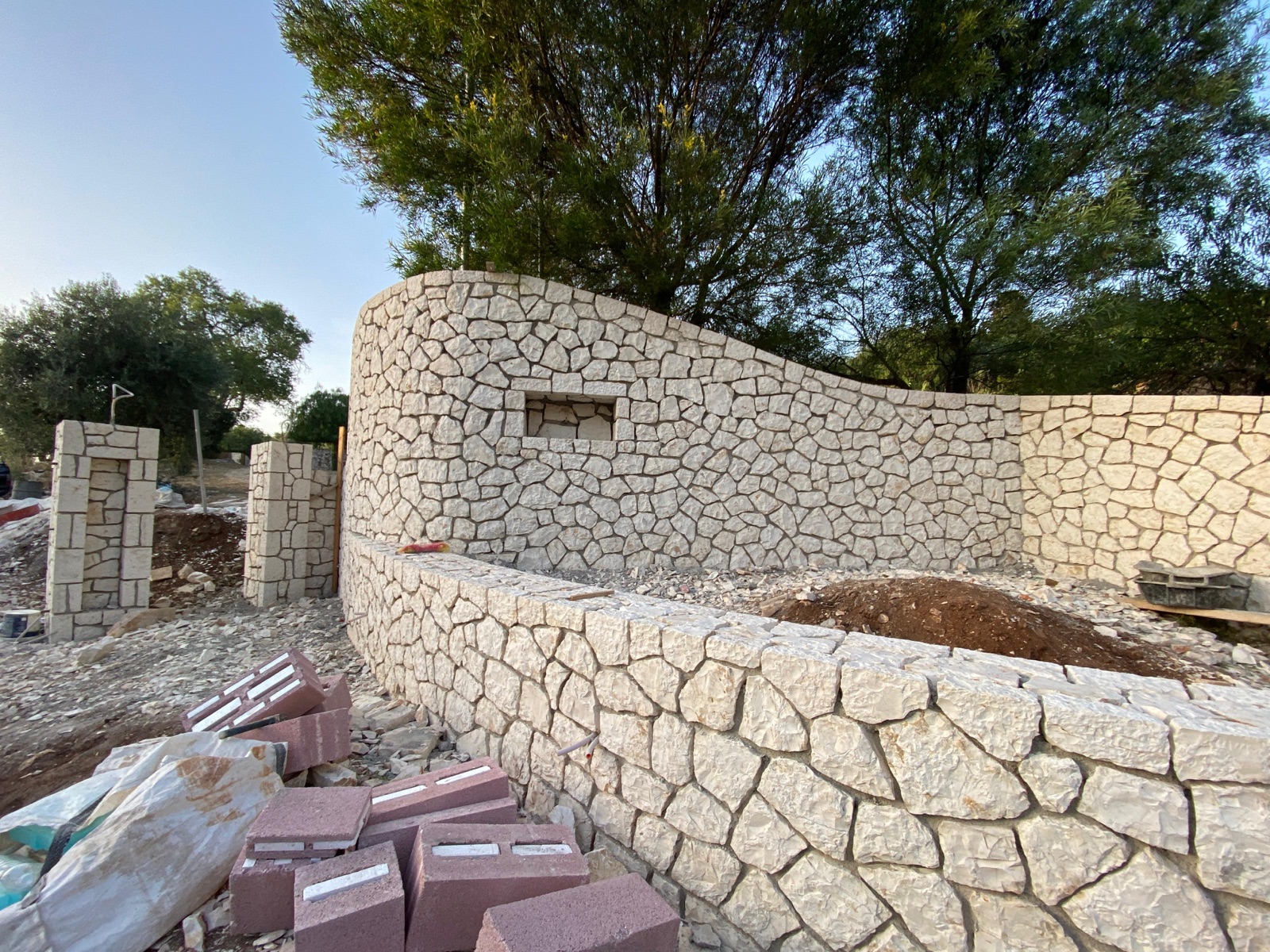 The serpentine cream limestone perimeter wall with built-in niche and gate pillars taking shape, building stone stacked in front.