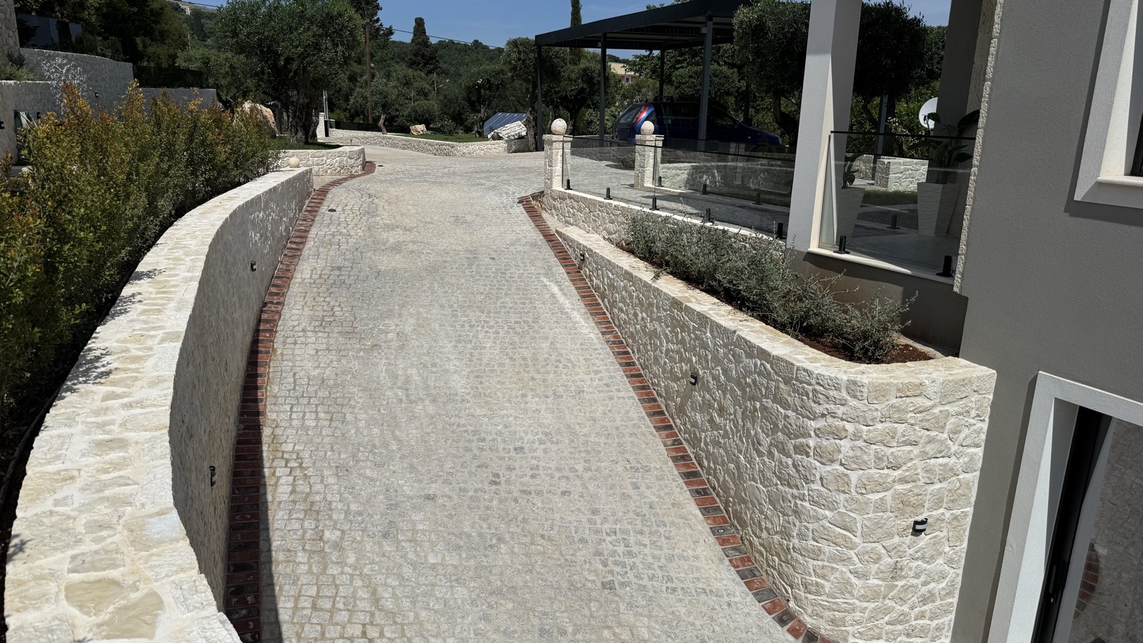 The cobblestone driveway curving between low white stone walls with red-brick trim, a planted stone planter wall alongside.
