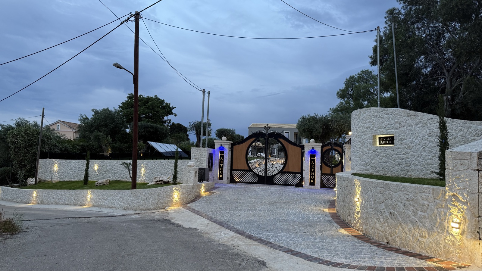 White stone perimeter walls washed by warm uplights at dusk, the ornate entrance gates lit between them.