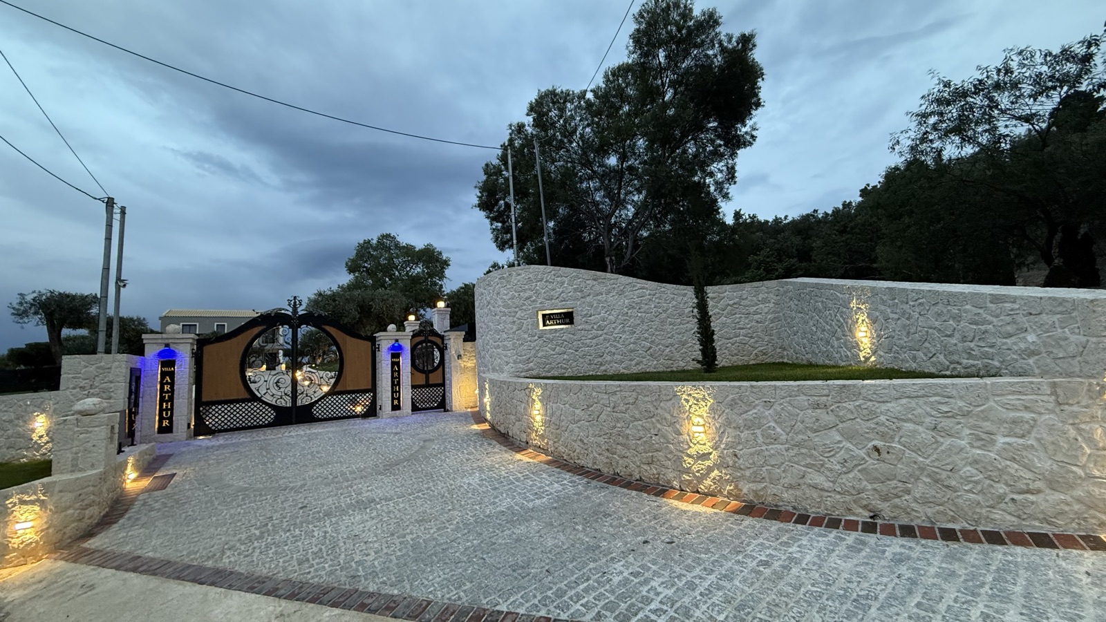 The gates at dusk with the serpentine stone wall sweeping right, warm sconces grazing the wall texture above the cobbled drive.