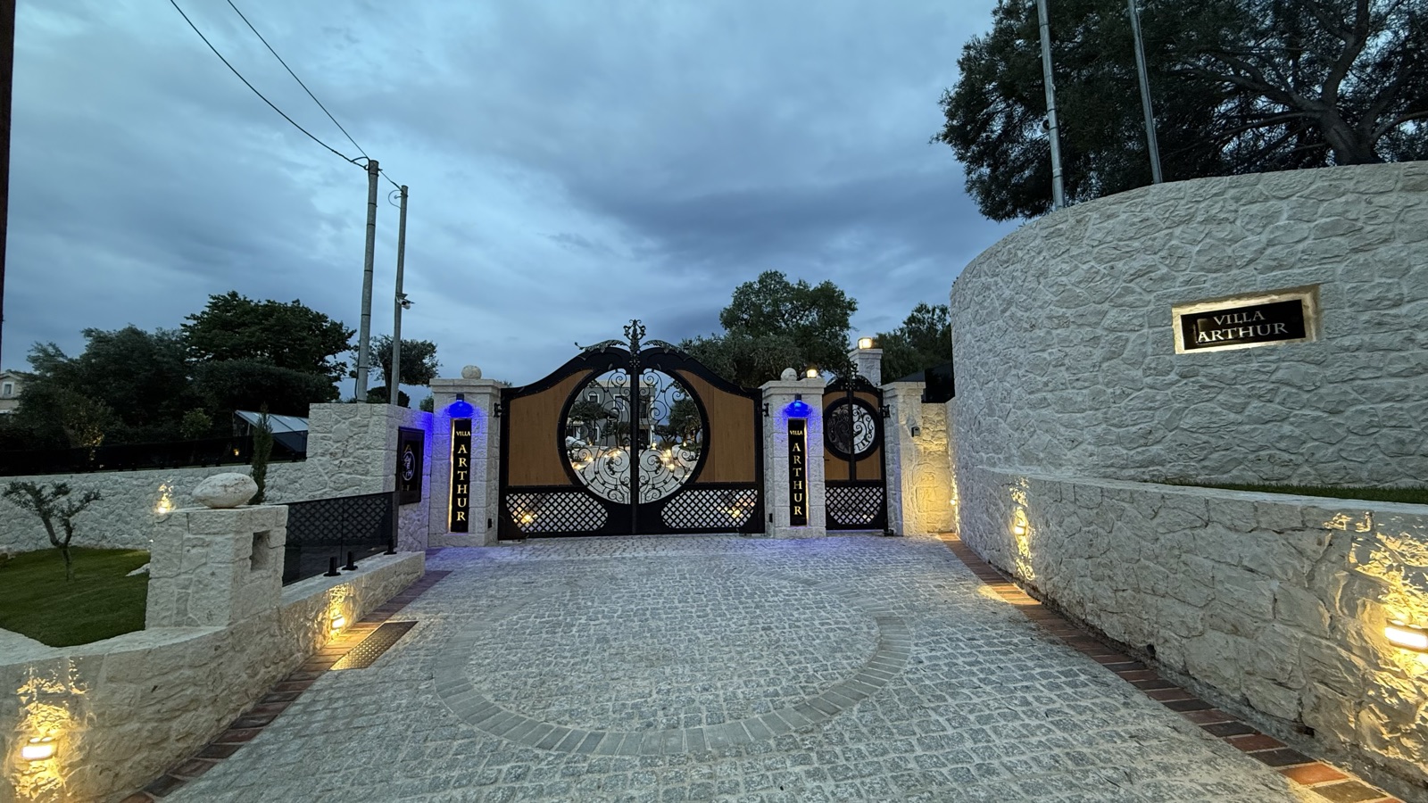 The closed ornate gates at dusk from inside the drive, a circular cobble pattern in the paving and warm light on the stone wall.