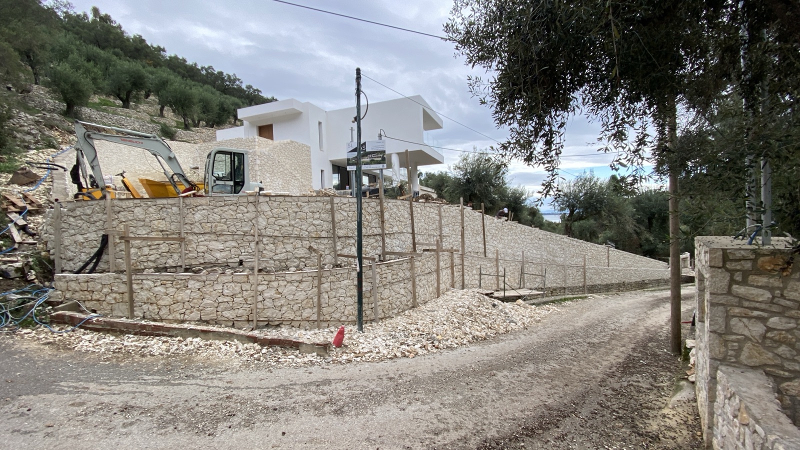 An excavator and mini-digger in front of new terraced dry-stone retaining walls, the white villa shell behind under a winter sky.