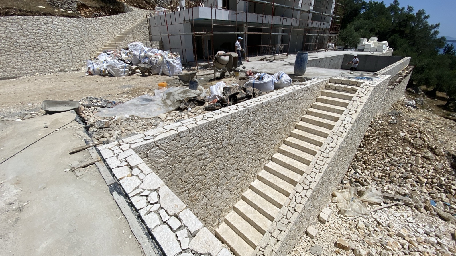 The stone-clad staircase and angled retaining walls taking shape beside the open pool shell, the sea in the distance.