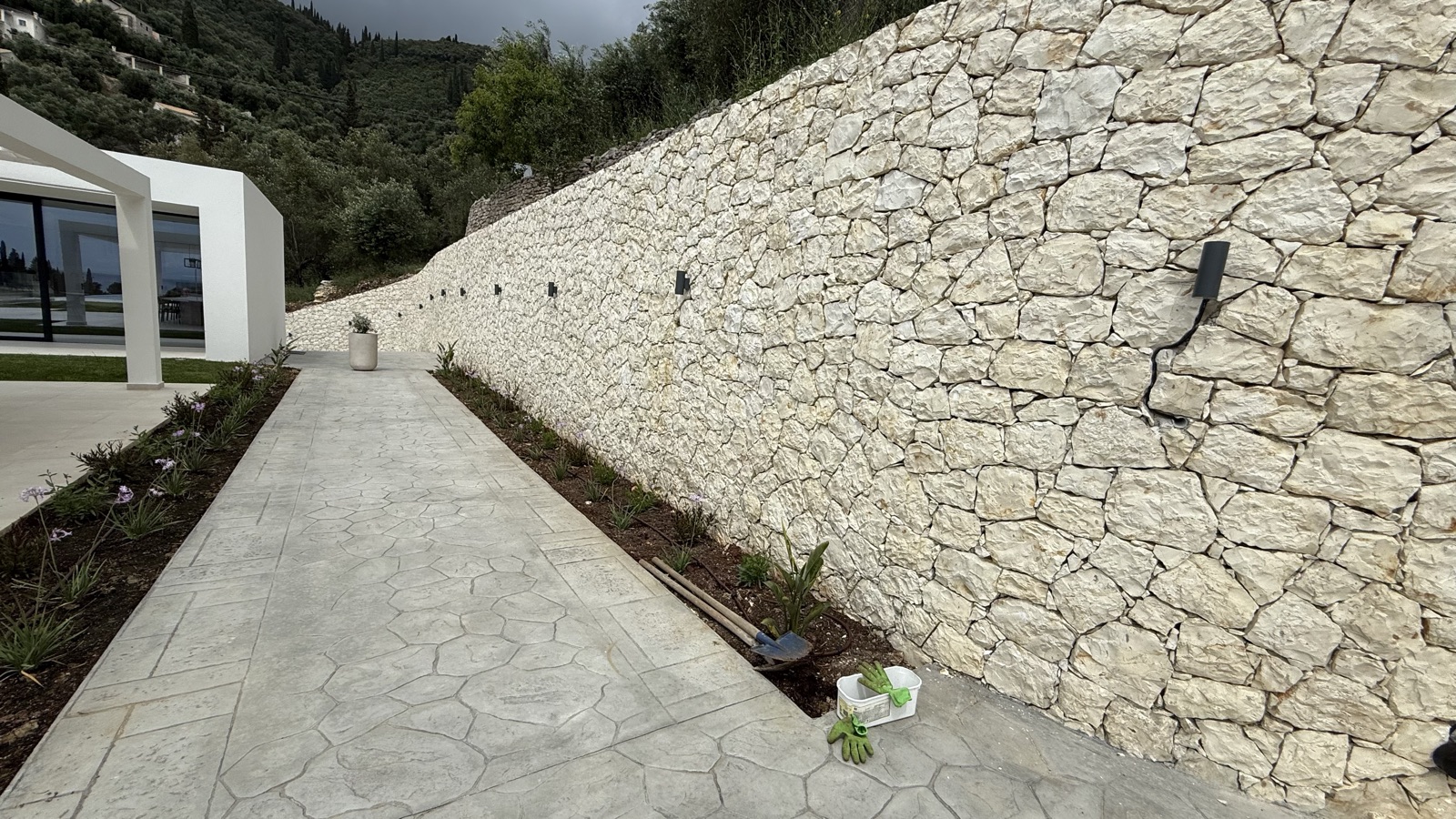A pathway running between the villa's white pergola and the long limestone wall, fresh planting beds along its base.