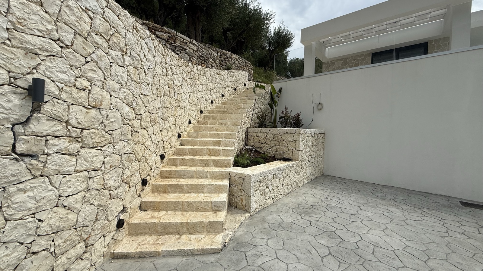 The limestone staircase climbing the rough-coursed stone wall to meet the white rendered villa, a raised planter at its base.