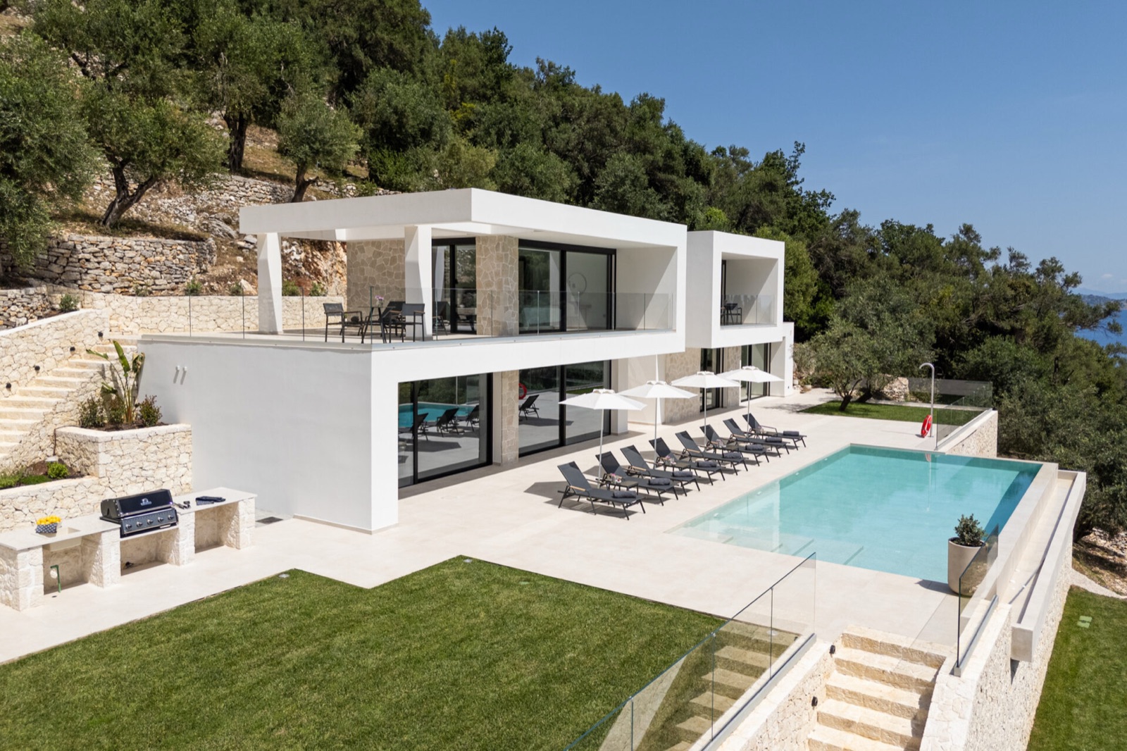 White cantilevered volumes on stone-clad piers above the infinity pool and lawn, terraced limestone walls and a glimpse of sea beyond.