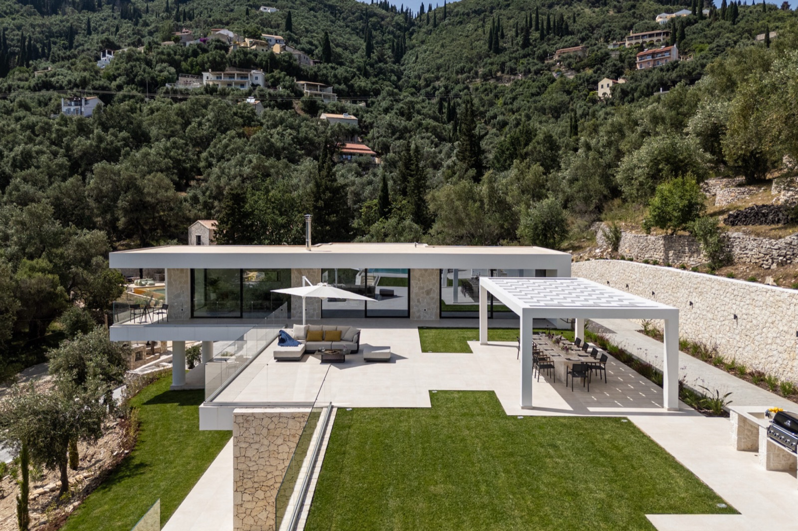 The flat-roofed living volume with stone-clad gable accent, a white pergola over the long dining table, curved limestone wall behind.