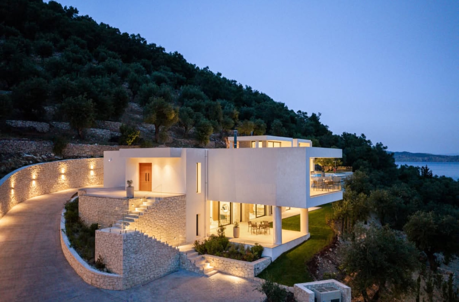 The villa glowing warm at dusk, limestone retaining walls uplit along the curving driveway, the sea and hills fading behind.