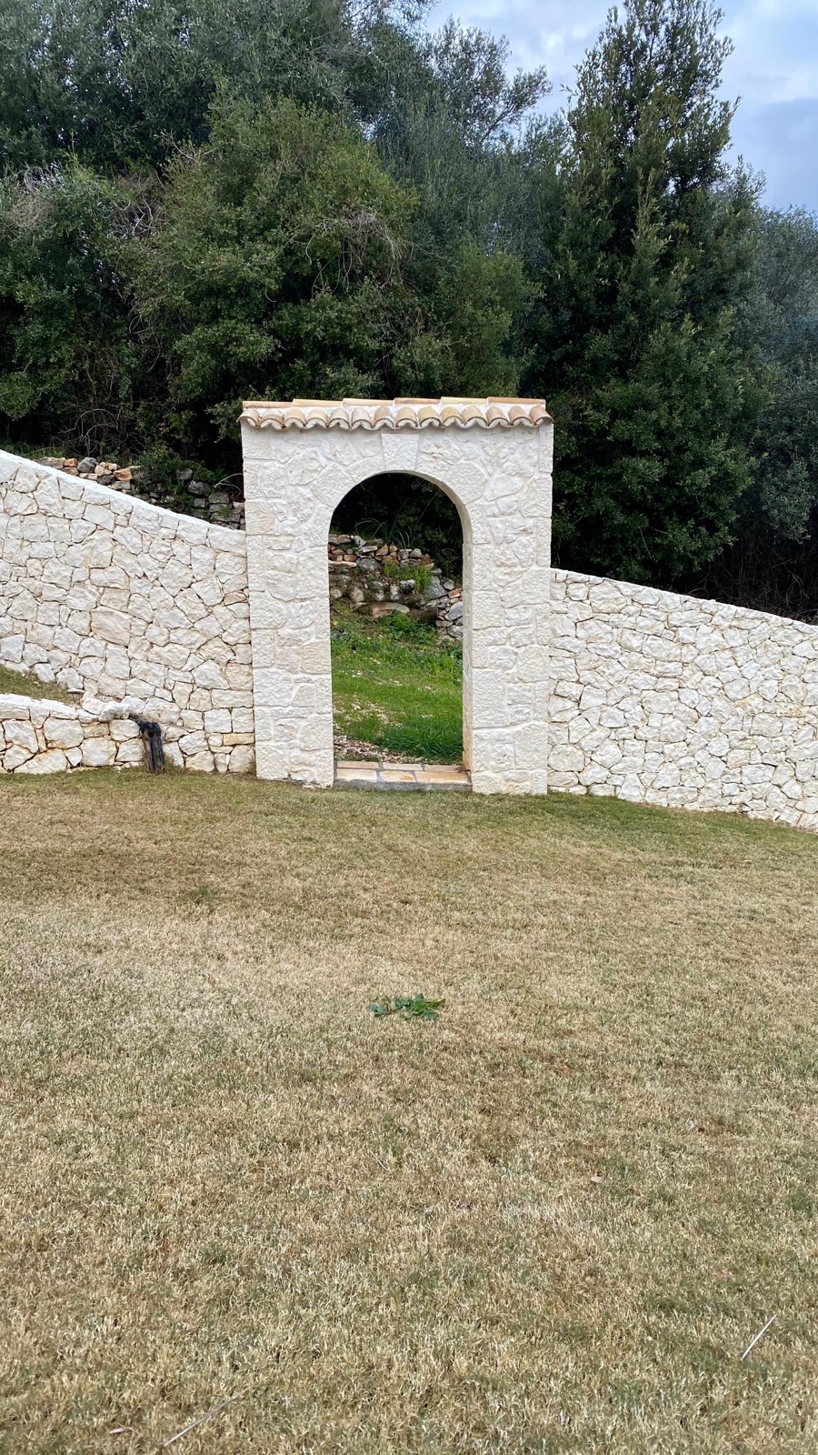 Freestanding arched stone gateway set into a serpentine cream stone wall capped with terracotta tiles, Villa No. 8, Corfu.