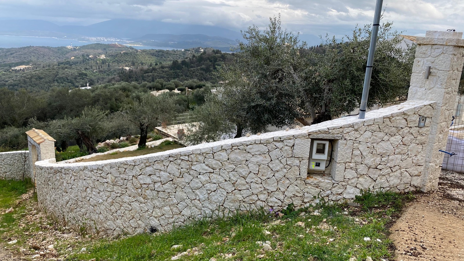 The sweeping curved cream stone perimeter wall and gate pillar, olive groves descending to the sea and mountains under moody clouds.