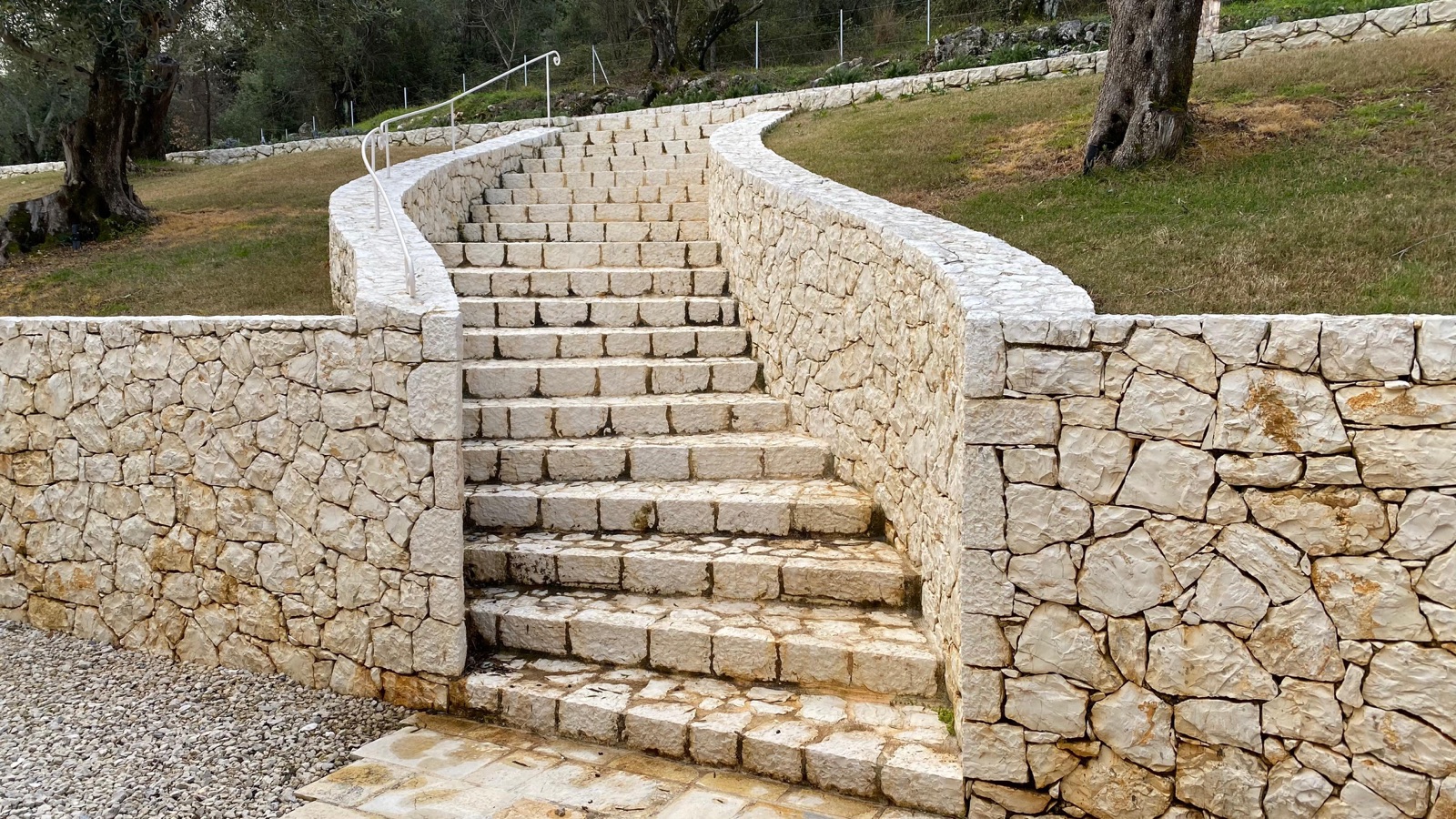 A curving rough-stone staircase flanked by serpentine cream walls with a slender wrought-iron handrail, olive trees and lawn above.