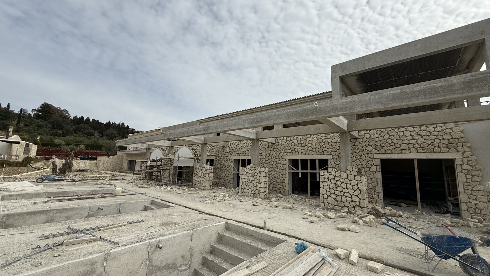 A row of round stone arches under white concrete pergola beams, with the raw pool shells open in the foreground.