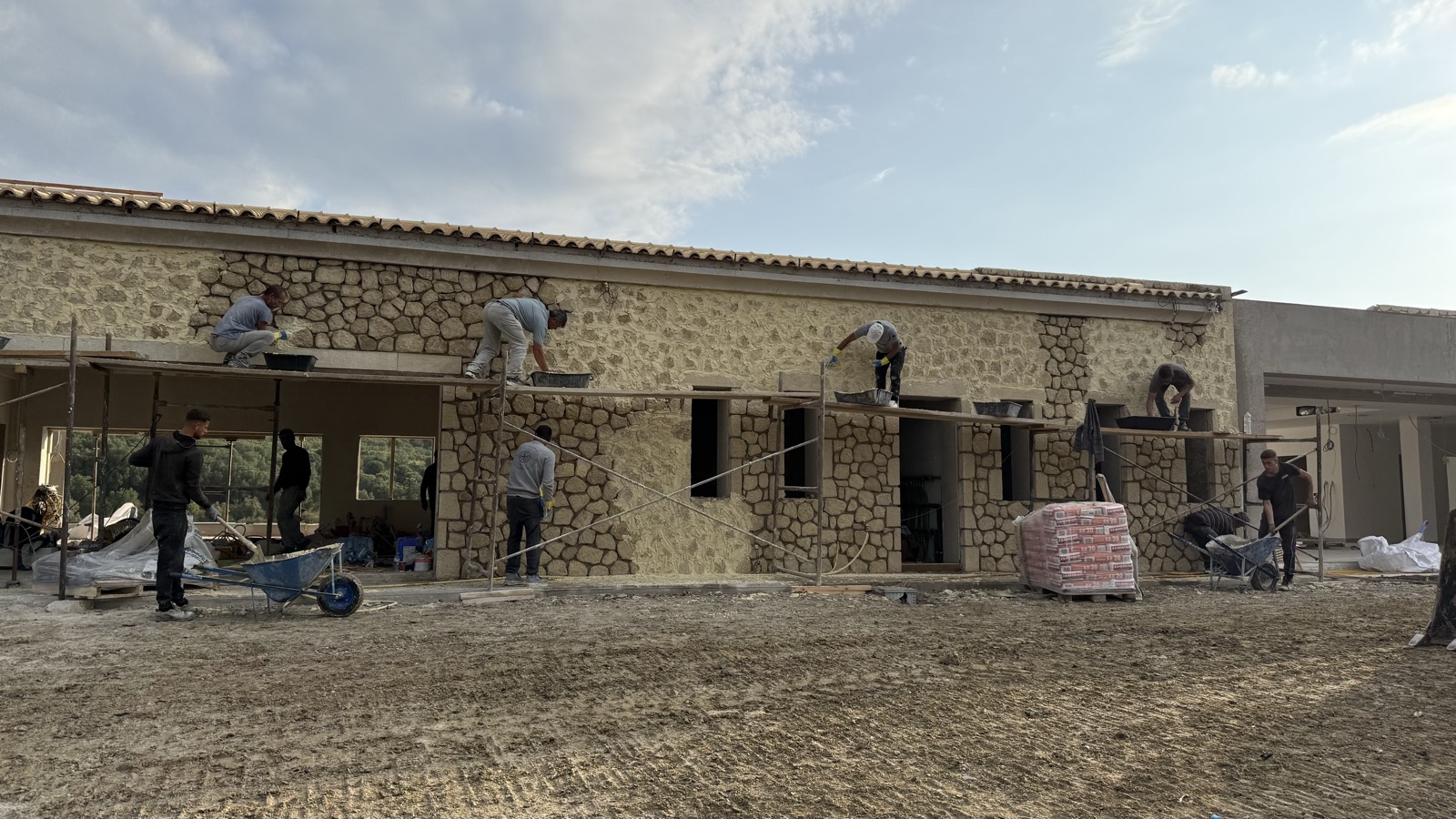 Masons on scaffolding laying fieldstone across the facade in warm low light, mortar trays and stone staged on the forecourt.