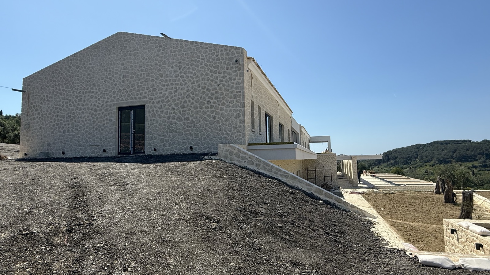 The stone gable end of the villa above the lower arcade, terraced planting beds with olive trees and wooded hills beyond.