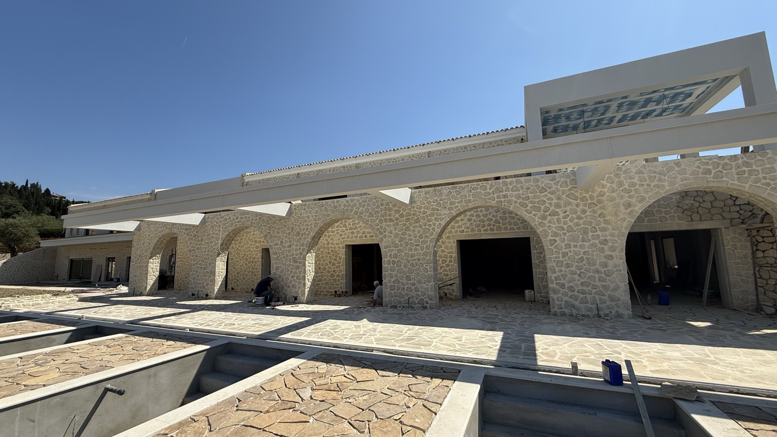 The full arcaded facade of repeating stone arches under a long white pergola, the flagstone terrace and open pool shells in front.