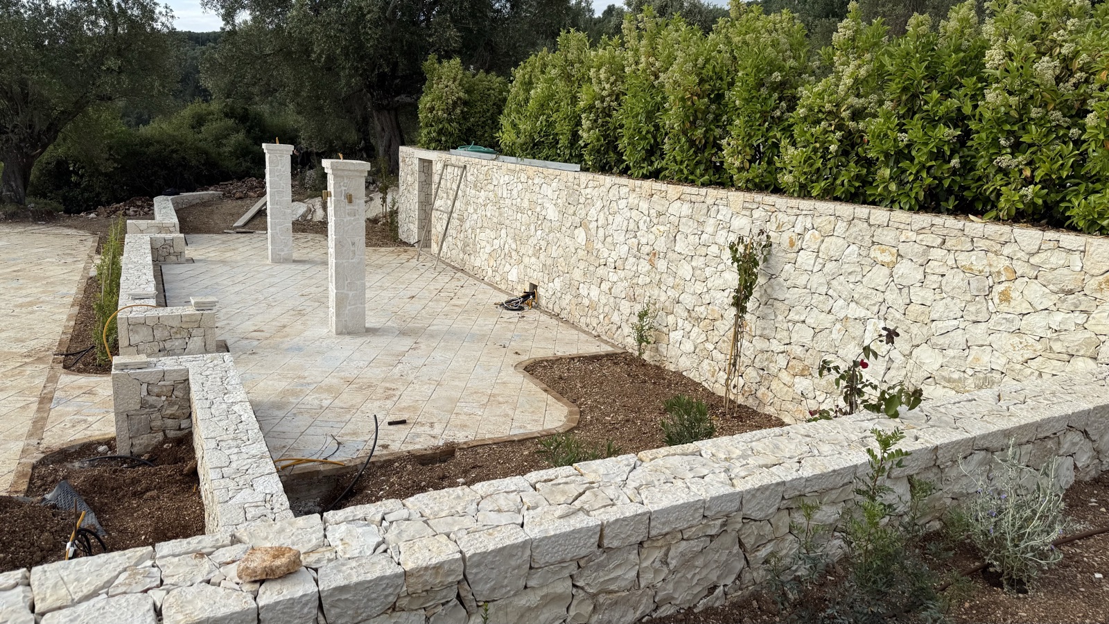 New pale dry-stack limestone garden walls and two square stone pillars, with fresh paving and young plantings by a green hedge.