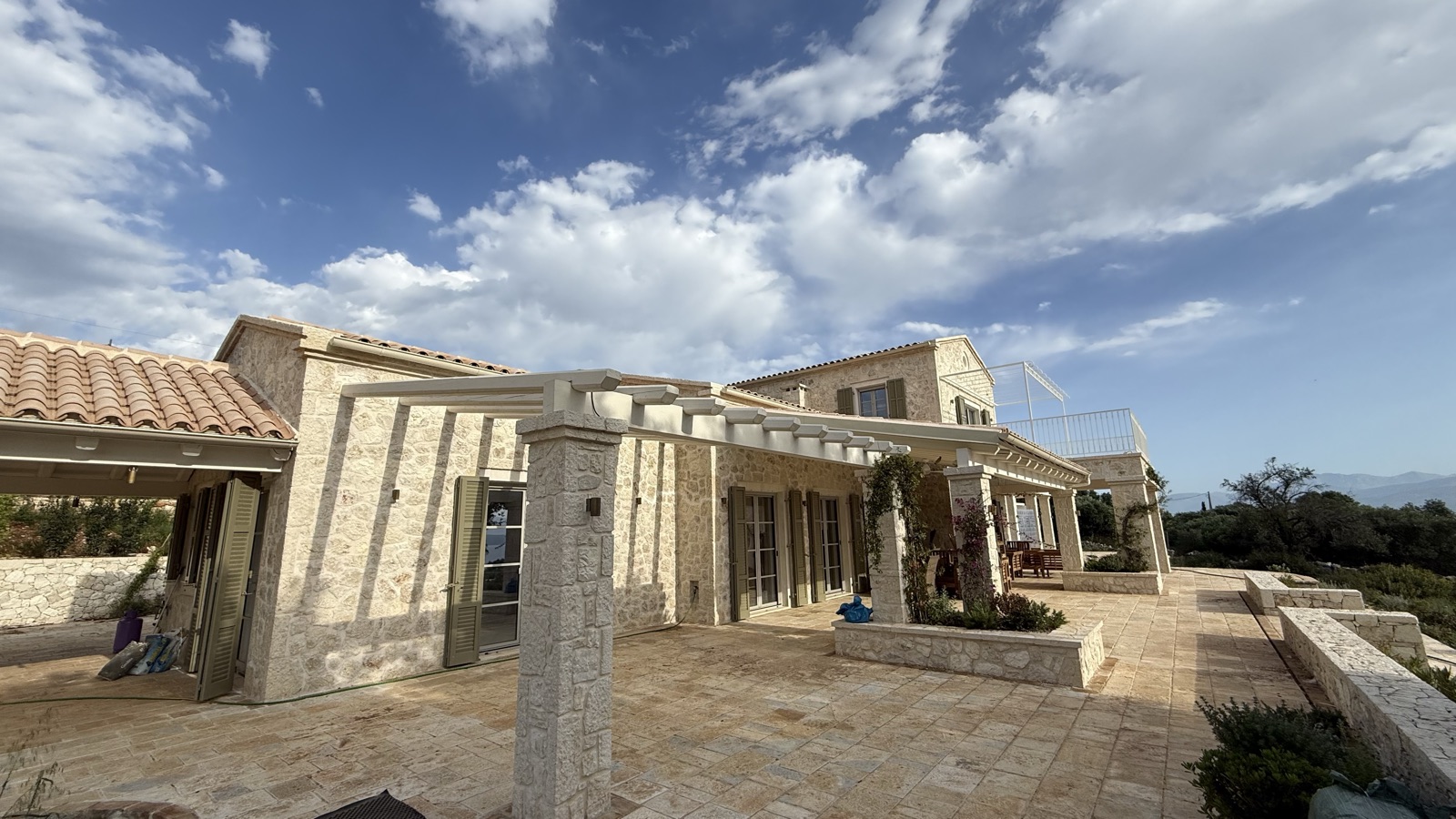 Stone-clad walls, white timber pergola on a stone column, sage shutters, terracotta roofline, dramatic blue sky with clouds