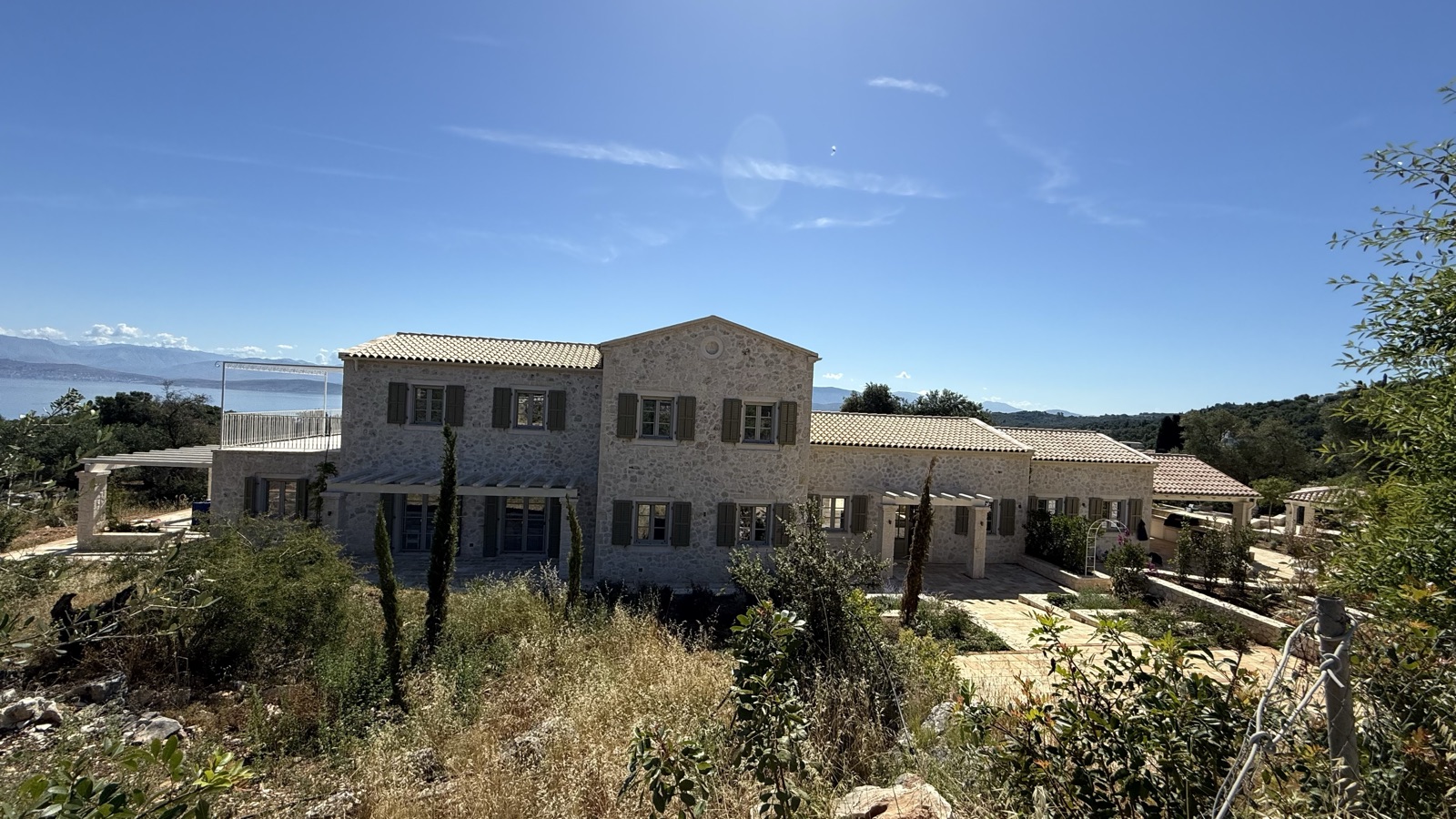 Full establishing shot of the entire two-story stone villa on its hilltop, sea and mainland mountains behind, clear blue sky, wild scrub in foreground