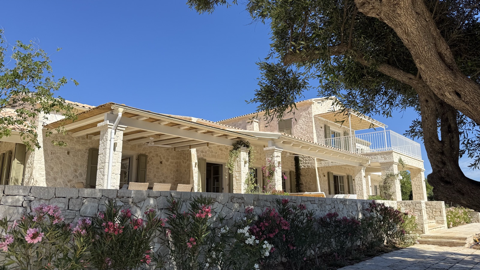 Finished facade with white pergola veranda seen over a dry-stone garden wall lined with pink and white oleander, olive tree canopy framing right, deep blue sky
