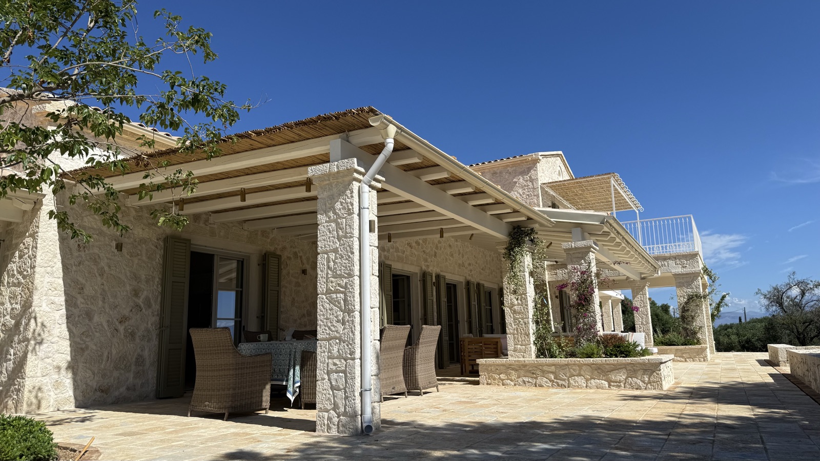 Rough stone columns, wicker dining chairs, climbing bougainvillea, upper balcony behind, blue sky