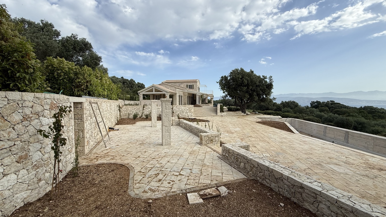 Stone retaining walls and pillars in the lower courtyard, the villa above and the sea and mountain horizon under dramatic clouds.
