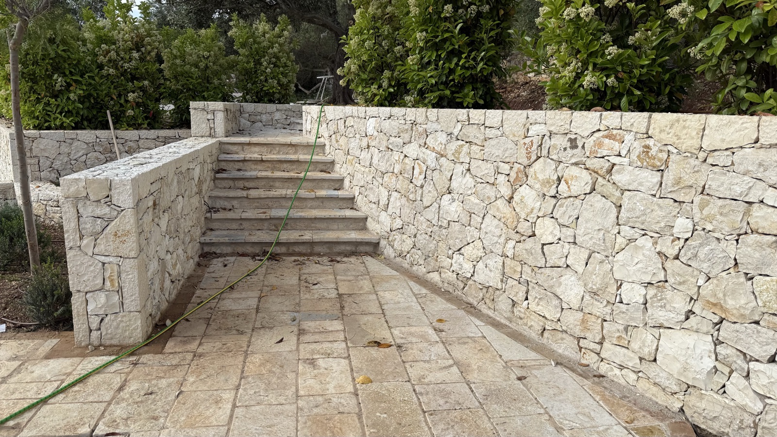 A pale rough-faced limestone wall and stone steps meeting cream flagstone paving, with greenery above.