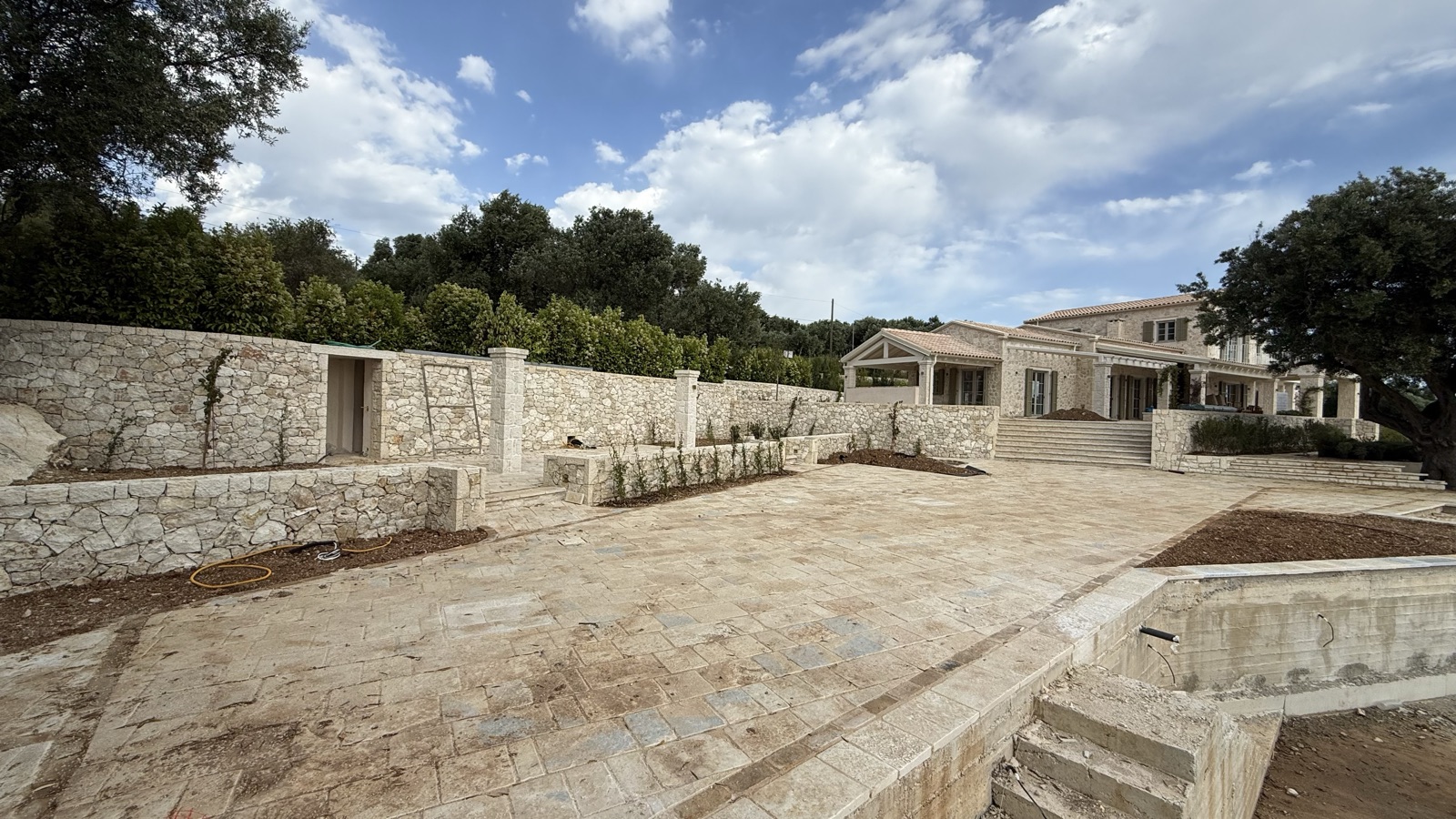 The long stone perimeter wall with its gate opening, planted beds and a large flagstone terrace looking back at the villa.