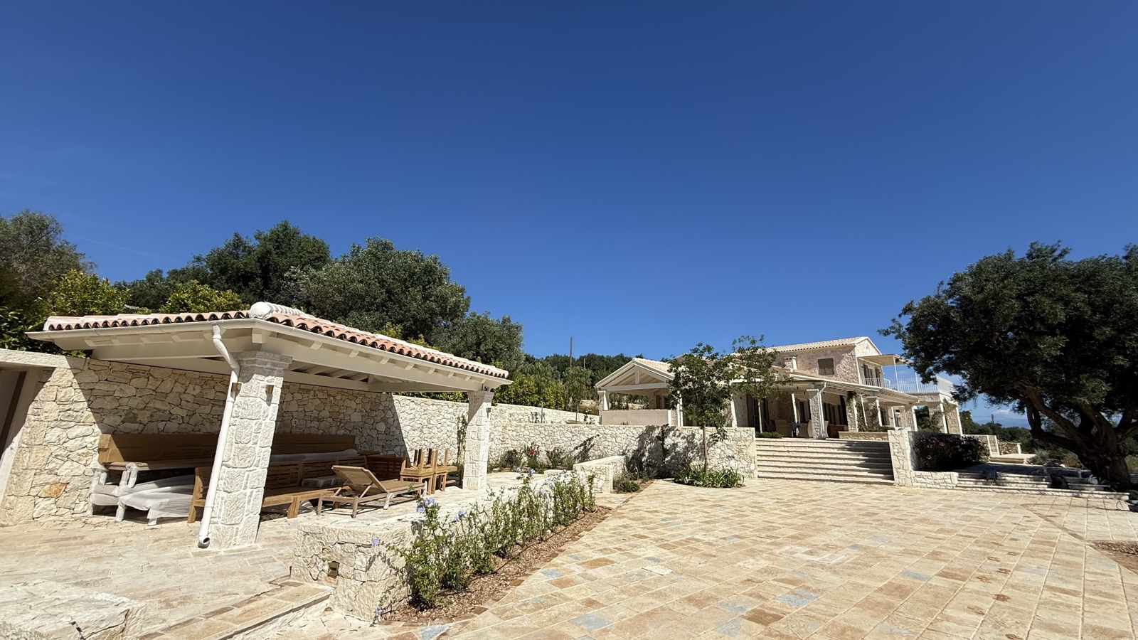 Stone pool pavilion with tiled roof and columns, loungers beneath, garden walls and villa in background, large tree right, deep blue sky