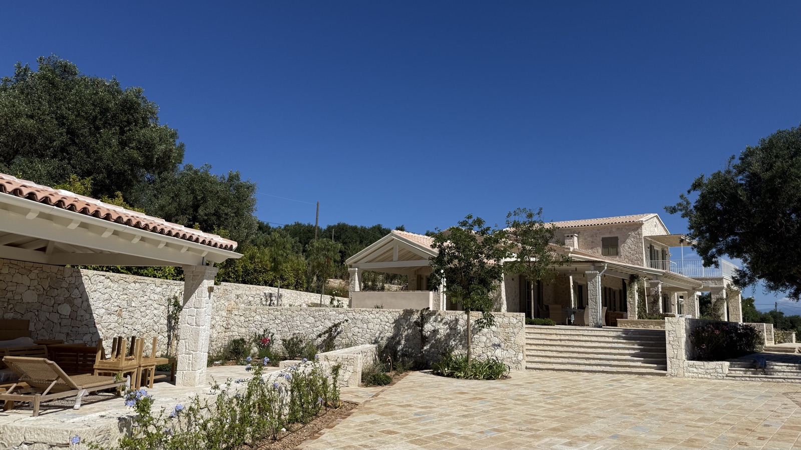 Pergola corner with wooden loungers, plumbago and flowering beds along stone walls, villa with grand steps in background, saturated blue sky