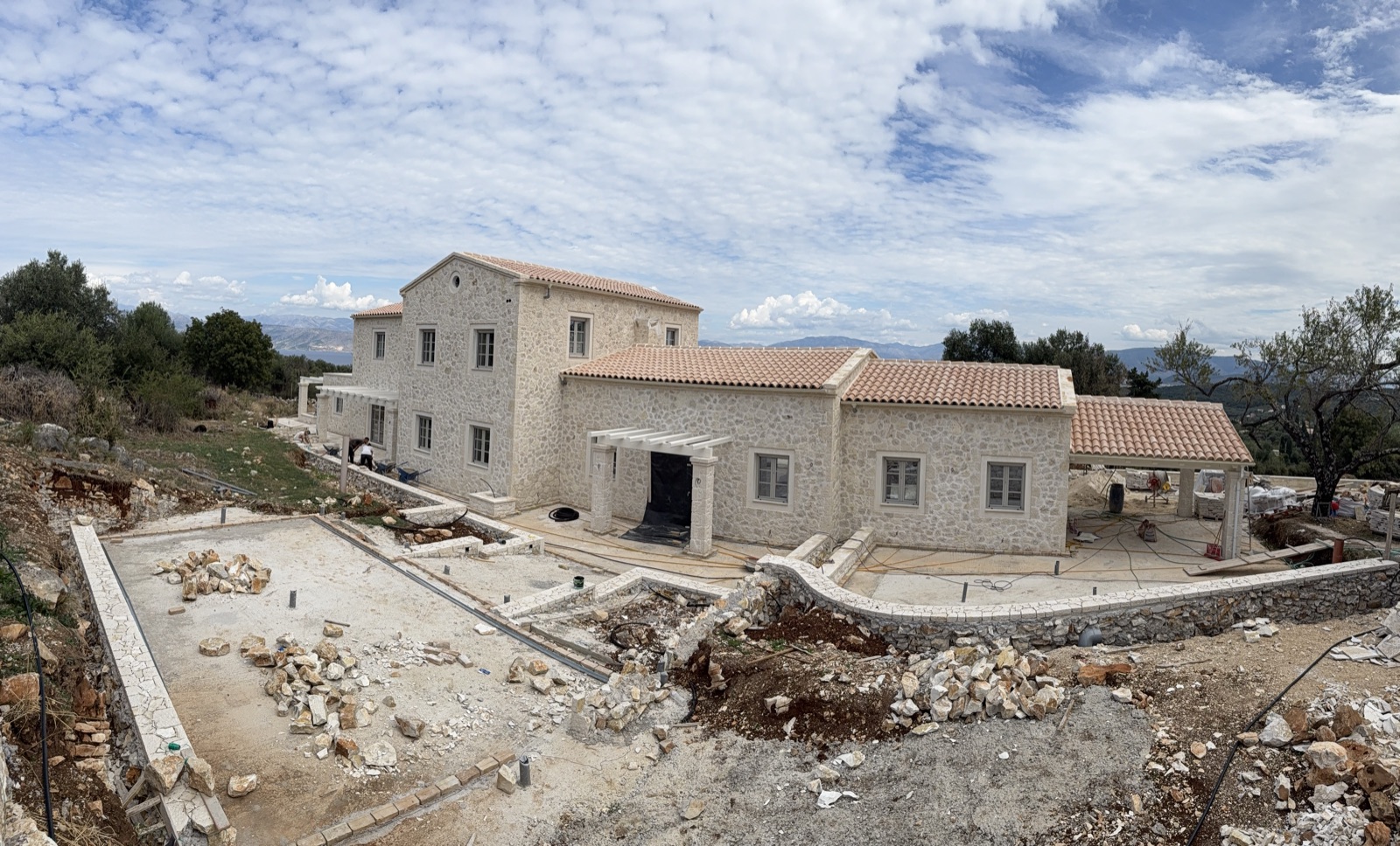 The stone villa standing complete above raw ground, stone heaps and unfinished walls all around under a brooding sky.