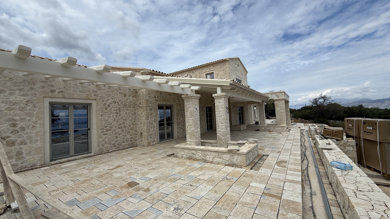 White pergola beams on stone columns over the completed mixed-stone paving, the two-storey villa behind.