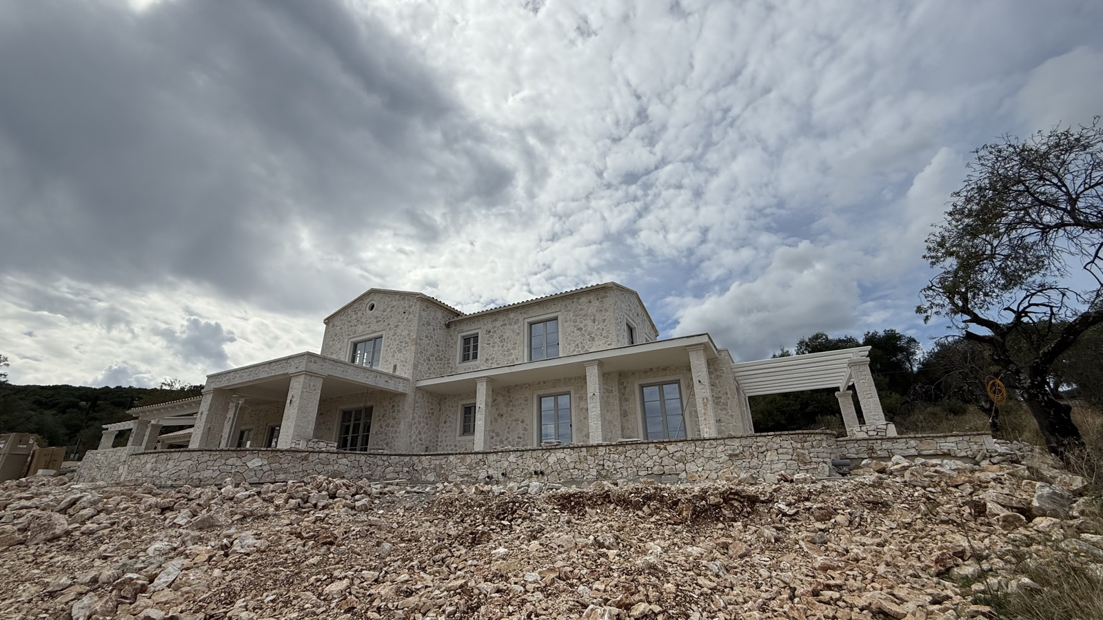 The full two-storey stone villa seen from the rocky slope below, under dramatic storm clouds.