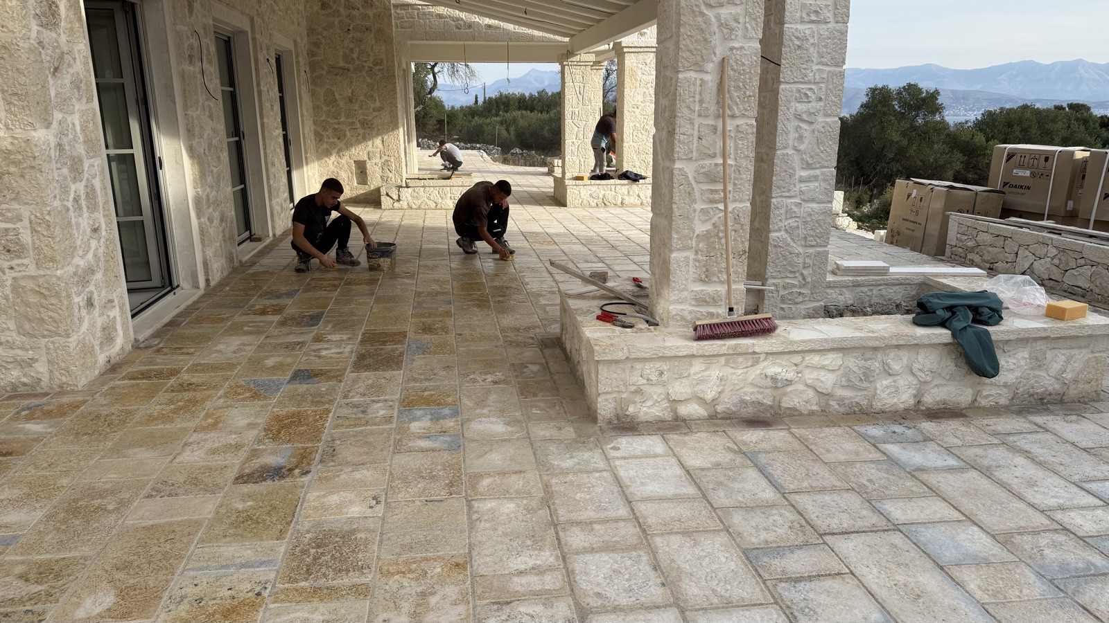 Three masons kneeling to grout the stone paving under the porch colonnade, with a glimpse of sea beyond.