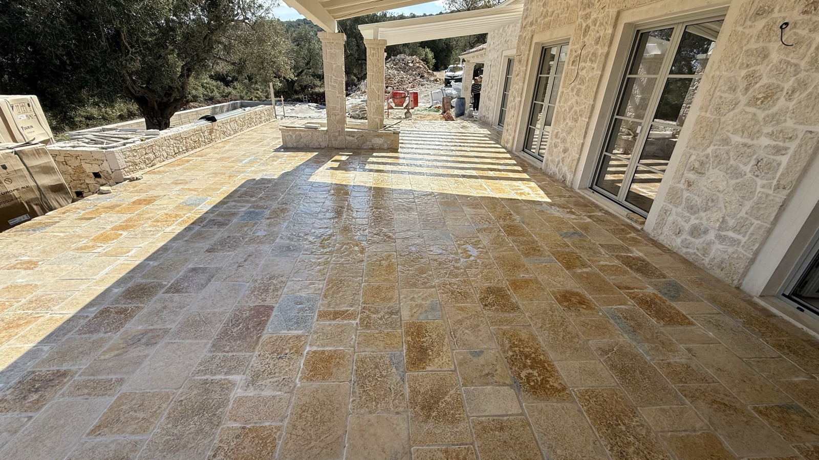 Freshly washed ochre-gold paving glowing wet across the main terrace, pergola shadows falling toward the stone facade.