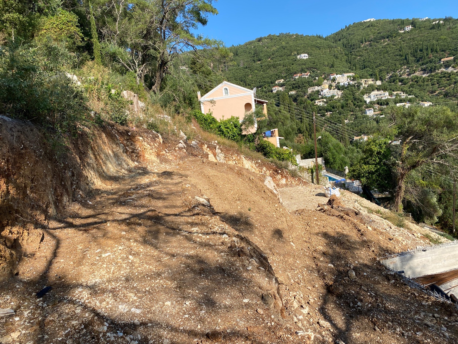 A graded earth terrace on the steep slope, with a neighbouring peach-coloured villa and the hillside village beyond.
